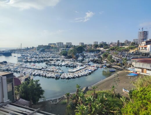 Vista panoramica del porto turistico di Baia di Ognina, Sicilia, con barche ormeggiate, spiaggia e pittoresco paesaggio urbano sullo sfondo.