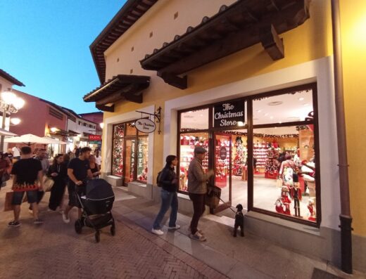 Barberino Outlet: People stroll past 'The Christmas Store' at dusk; shop windows display festive decorations, attracting shoppers and their small dog.