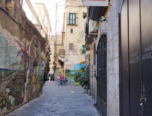 Suggestiva veduta di una stretta via a Napoli con un murales colorato e persone sedute a tavoli all'aperto; ideale per esplorare i viaggi e la cultura locale con Barbirotti Viaggi dal 1913