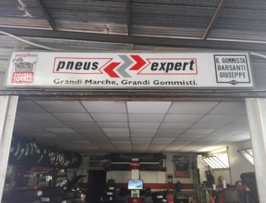 L'insegna di "Pneus Expert" di Giuseppe Barsanti, un gommista specializzato in grandi marche, con garanzia totale. Interno dell'officina con pneumatici e attrezzature visibili.