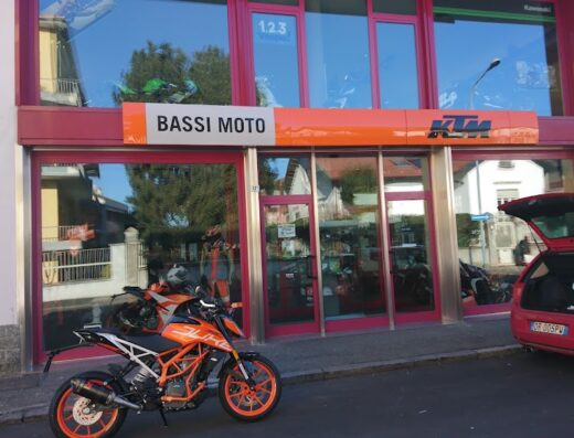 Bassi Moto concessionaria KTM: moto arancione Duke parcheggiata di fronte al negozio, auto rossa con portellone aperto, accessori e caschi visibili.