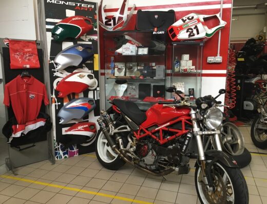 Esposizione di una moto Ducati rossa presso BAT Moto Service, con serbatoi colorati, accessori Ducati e abbigliamento da motociclista.