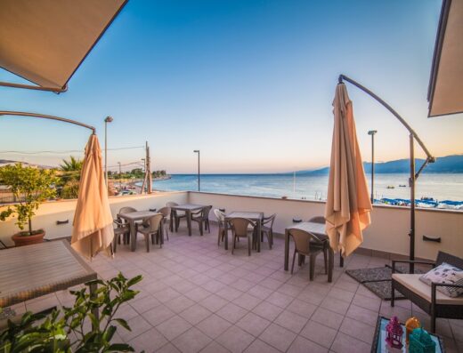 Terrazza panoramica di B&B a due passi dal mare con tavoli, sedie e ombrelloni chiusi, vista sulla costa e sul mare al tramonto.