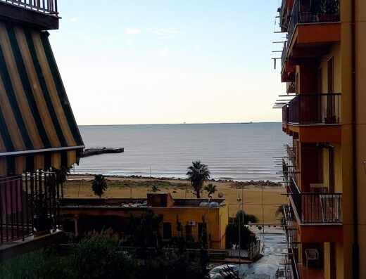 Vista mare da B&B a Gela: spiaggia sabbiosa, palme, edifici colorati e mare calmo all'orizzonte.