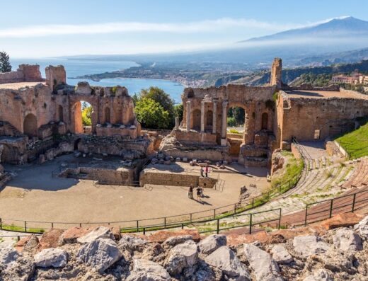 Veduta panoramica del Teatro Antico di Taormina, Sicilia, con l'Etna sullo sfondo e turisti che visitano l'attrazione.
