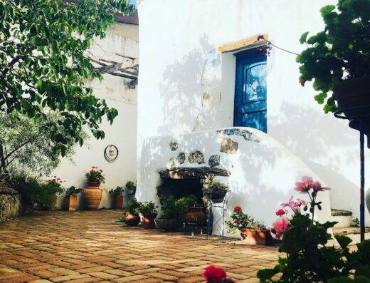 Corte interna di B&B a Oliena, Sardegna, con pavimentazione in cotto, fiori in vaso, pareti bianche e porta blu.