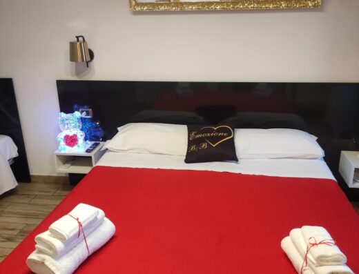 Camera da letto accogliente di B&B Emozione con letto matrimoniale coperto da un lenzuolo rosso, cuscino decorativo e asciugamani bianchi piegati.