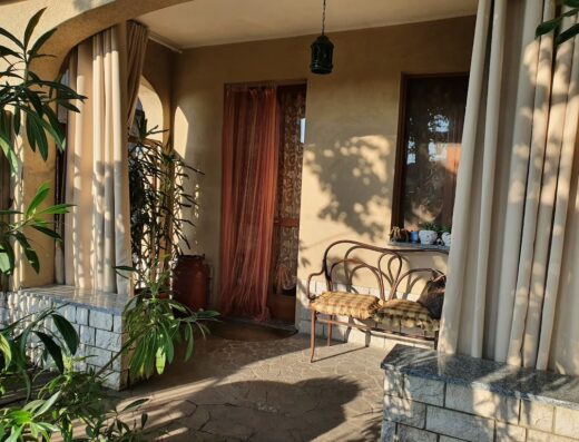 Veranda di una casa di famiglia con tendaggi beige, una panca in ferro battuto con cuscini, piante in vaso e una lanterna sospesa.