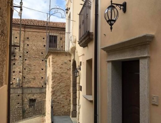 Stretta via del borgo antico, con edifici in pietra e intonaco chiaro, balconcini in ferro battuto e lampade decorative.