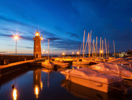 Il porto turistico di Desenzano del Garda al crepuscolo, con il faro illuminato e le barche ormeggiate, riflessi nell'acqua calma.