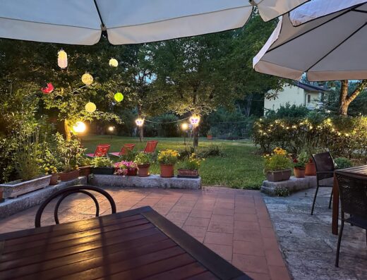 Patio esterno illuminato di sera con tavolo in legno, sedie, ombrelloni, fiori in vaso e lanterne decorative sospese tra gli alberi.