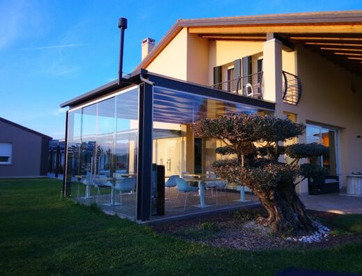 Veranda in vetro di B&B Magrine con tavoli e sedie, illuminata dalla luce del sole al tramonto, accanto a un ulivo ornamentale.
