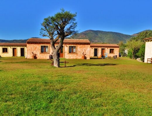 B&B Pubulos: Vista di un prato verde con case vacanza in stile rustico, caratterizzate da tetti rossi e pareti color terra, sotto un cielo azzurro.