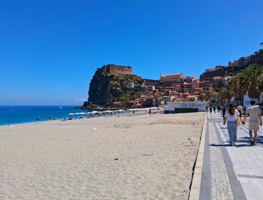 Veduta panoramica di Scilla, Calabria, con spiaggia assolata, mare cristallino, borgo storico sul promontorio e persone che passeggiano sul lungomare.