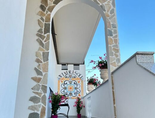 Ingresso del B&B Via del Sole con arco in pietra, decorazioni floreali e insegna con il nome, un'accogliente struttura ricettiva per viaggiatori in moto.
