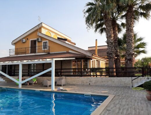 B&B Vittoria House exterior view with swimming pool, palm trees, outdoor seating, and two-story building, showcasing the B&B's relaxing atmosphere and architecture.