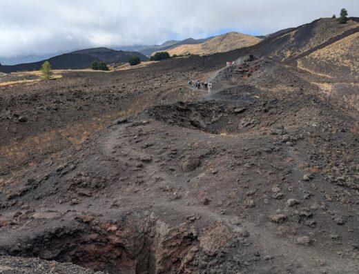 Escursionisti percorrono un sentiero su un terreno vulcanico con un cratere in primo piano, paesaggio di viaggio in Sicilia.