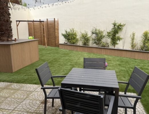 Cortile esterno arredato con tavolo e sedie in metallo nero, prato verde curato, illuminazione festosa e vegetazione decorativa sullo sfondo.