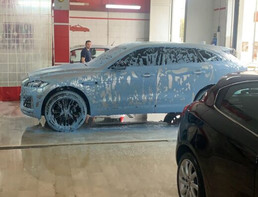 Auto di lusso bianca ricoperta di schiuma durante un lavaggio auto presso Beauty Car Wash, con un addetto ai lavori visibile sullo sfondo.
