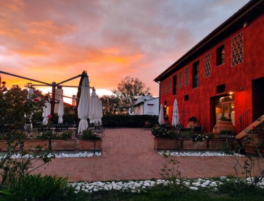 Borgo San Giusto Bed and Breakfast exterior view at sunset, showcasing the red building, outdoor seating with umbrellas, and a tranquil courtyard in Tuscany, Italy.