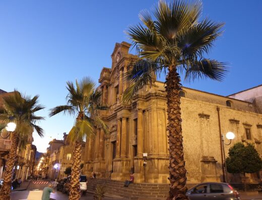 La facciata barocca di una chiesa a Piazza Armerina, Sicilia, con palme e cielo sereno al tramonto. Location ideale vicino al Bed and Breakfast La Volpe Rossa.