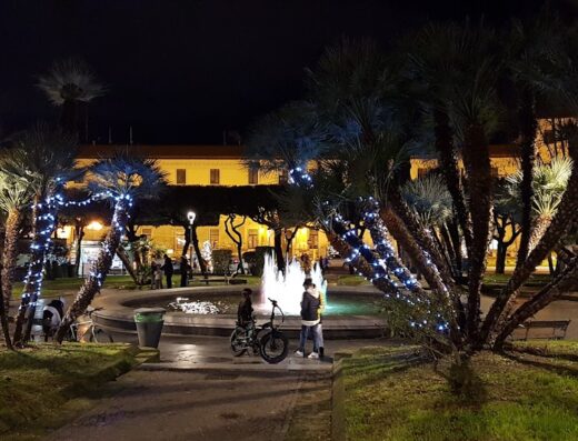 Bed & Breakfast Le Fontane del Re: A nighttime view of a fountain surrounded by palm trees adorned with lights, with people and a motorcycle nearby.