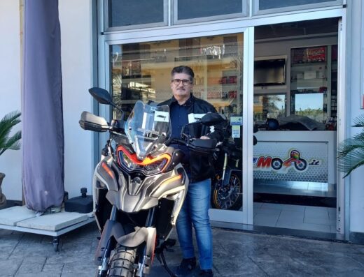 Un uomo in piedi accanto a una moto adventure di fronte a un negozio di moto Benny Moto, con insegna Piaggio visibile.