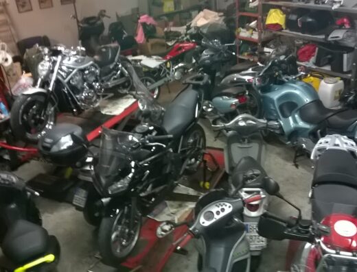 Officina moto affollata con diverse motociclette e scooter in riparazione, accessori moto e attrezzature da officina visibili, ambiente di lavoro per la manutenzione moto.