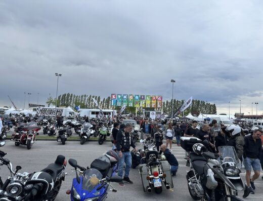 Affollato parcheggio moto al Biker Fest International, con diverse marche e modelli. Accessori moto e stand visibili sullo sfondo.