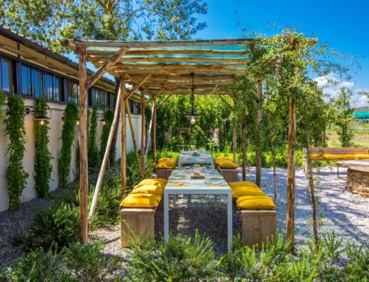 Area pranzo esterna con pergola in legno al Bio Agriturismo Il Cerreto, tavoli imbanditi e cuscini gialli per un'esperienza culinaria immersa nella natura.
