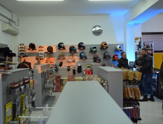 Interni del negozio BisonMotorParts con esposizione di caschi moto, occhiali, guanti e accessori per motociclisti. Clienti che acquistano ricambi e abbigliamento tecnico.