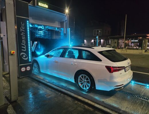 Un'Audi A6 bianca station wagon in un autolavaggio WashTec con illuminazione blu, durante la notte, presso il punto vendita IP in via del Centenario 6A.