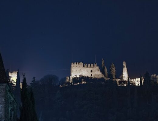 Borgo Castello illuminato di notte, con la torre di una chiesa in primo piano e il castello medievale sulla collina sullo sfondo.