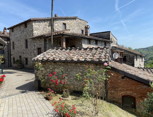 Borgo Giusto in Toscana: vista esterna delle case in pietra con tetti rossi, giardino con rose rosse e cielo azzurro.