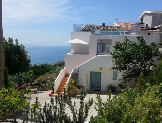 Borgo Rosso di Sera: Vista di una casa bianca con scale arancioni, ombrellone e vista mare, immersa nel verde.