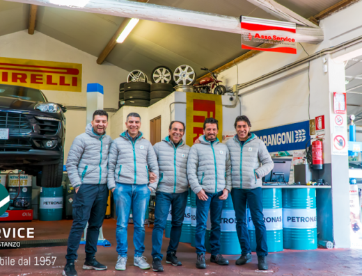 Foto di gruppo dello staff di Costanzo Car Service, officina Bosch Car Service dal 1957, con auto sul ponte sollevatore, pneumatici Pirelli e moto d'epoca sullo sfondo.
