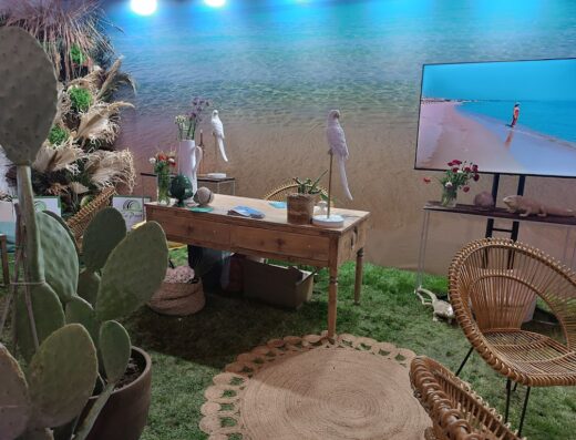 A decorative booth for BTM Puglia featuring a cactus, a wooden table with flowers and bird figures, a beach scene backdrop, and rattan chairs.