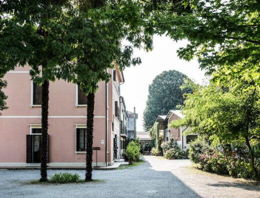 Ca' de Memi property: A charming pink building with dark shutters, framed by lush trees and a gravel driveway, creating a serene and inviting atmosphere.