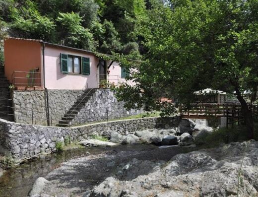 Cà du Ferrà Farm & Relax: Vista di un edificio rosa con persiane verdi affacciato su un ruscello con un ponte di legno in Liguria, Italia.