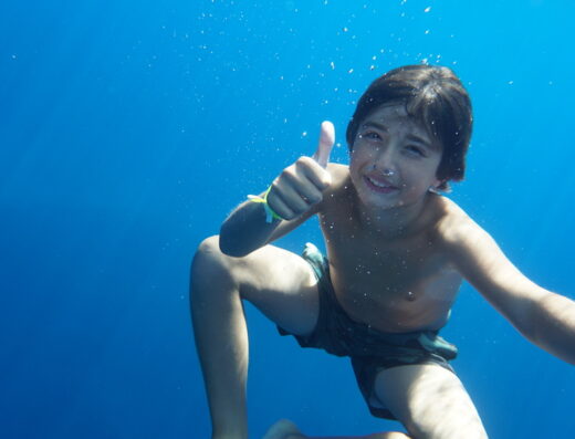 Ragazzo sorridente che fa il pollice in su sott'acqua con un'imbarcazione visibile in superficie, a Cagliari. Servizi di noleggio e turismo.