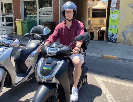 Un giovane uomo con casco guida uno scooter nero a Cagliari, Sardegna, di fronte a un negozio di noleggio moto. Scooter disponibile per esplorare la città.