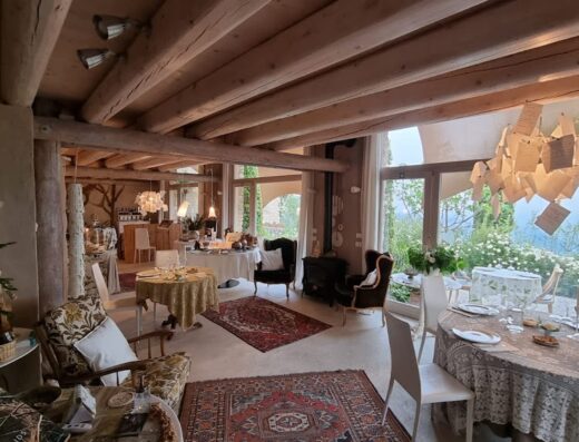 Interno di Calvenere con tavoli apparecchiati, sedie in stile, tappeti persiani, travi in legno a vista, lampadario di design e vista panoramica sul paesaggio circostante.
