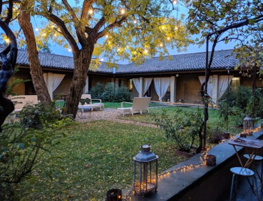Cortile interno del Cappuccini Resort con prato verde, sedie bianche, alberi illuminati e luci decorative, creando un'atmosfera accogliente.