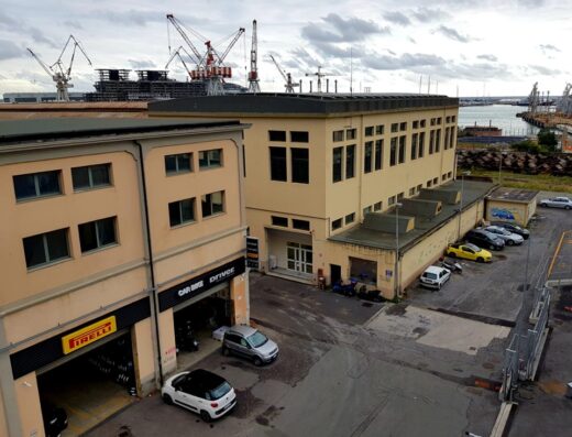 Esterno del Driver Center Pirelli, specializzato in auto e moto, con gomme Pirelli in esposizione e auto parcheggiate. Sullo sfondo, gru portuali.