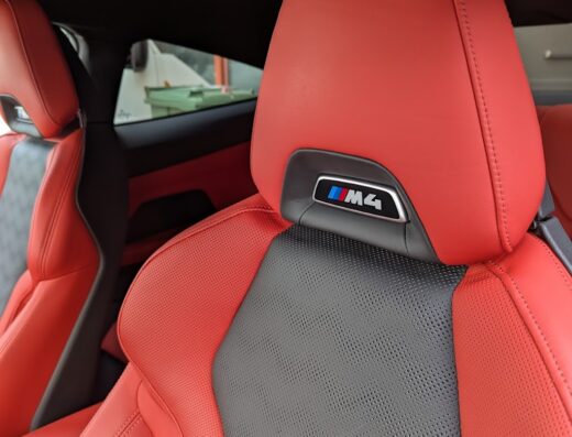 Primo piano dei sedili in pelle rossa e grigia di una BMW M4 con il logo M4 ben visibile sul poggiatesta. Interni auto di lusso.