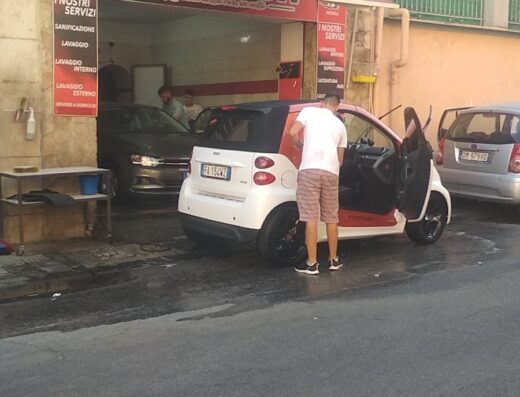 A white and red Smart car is being detailed at a car wash, with a man standing by the open door, and other cars are visible in the background.