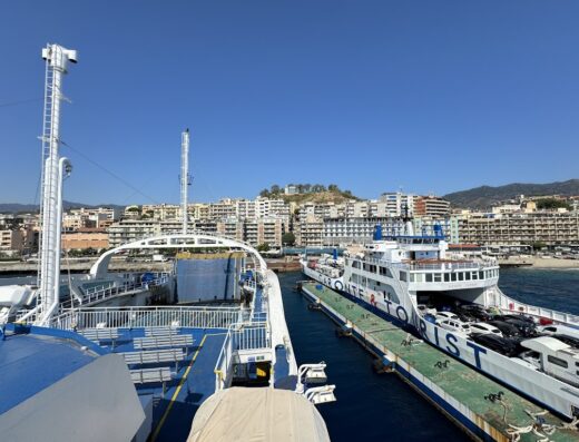 Vista ravvicinata dei traghetti Caronte & Tourist, con auto a bordo, attraccati in porto, sullo sfondo edifici della città.