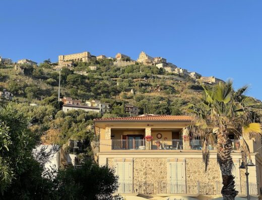 Affascinante vista di Casa del Sole, una residenza con balcone fiorito e facciata in pietra, sullo sfondo una collina dominata da antichi edifici.