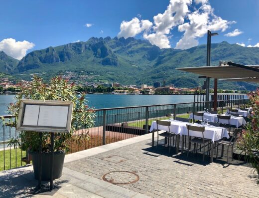 Ristorante con vista panoramica sul lago di Como, con tavoli apparecchiati all'aperto e menu esposto, sullo sfondo le montagne.