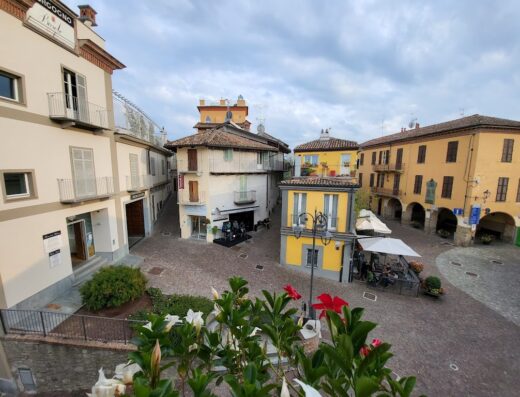 Vista panoramica di Barolo, Italia, con piazza centrale, architettura storica, bar con tavolini all'aperto e vegetazione in primo piano, ideale per agriturismo e turismo.
